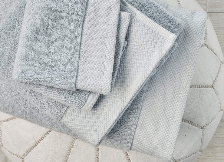 Stacked set of envello light blue cotton Washcloths and Bath Towels on a white leather pouf